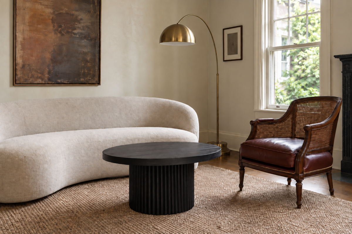 A curved contemporary sofa and an oxblood leather cane-back chair share the same brass and ebonized-oak thread in a plaster-walled living room.