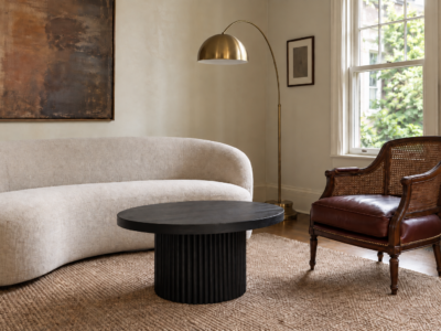 A curved contemporary sofa and an oxblood leather cane-back chair share the same brass and ebonized-oak thread in a plaster-walled living room.