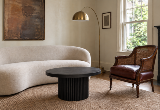 A curved contemporary sofa and an oxblood leather cane-back chair share the same brass and ebonized-oak thread in a plaster-walled living room.