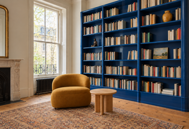 Built-in bookshelves with a glossy cobalt back, full of books and a few objects, beside an ochre lounge chair.