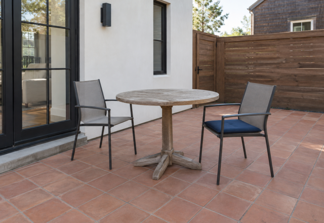 A small patio with a round teak pedestal table and two sling chairs, sized to leave clear space around it.