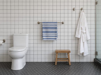 A bathroom wall of white square tile with a paper holder, a towel bar, and robe hooks mounted at different heights.