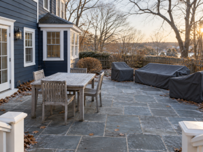 A patio beside a navy clapboard house on a frosty morning, with a bare teak dining set and a sofa set under fitted covers.