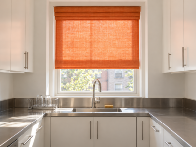 A tangerine linen roman shade glowing with daylight over the stainless sink of a white kitchen.