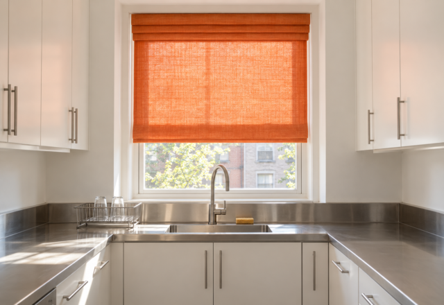 A tangerine linen roman shade glowing with daylight over the stainless sink of a white kitchen.