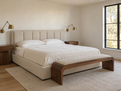 A primary bedroom with a paneled boucle headboard, brass wall sconces, and a curved wood bench at the foot of a low platform bed, morning light from a black-framed window.