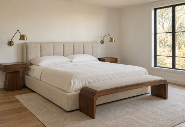 A primary bedroom with a paneled boucle headboard, brass wall sconces, and a curved wood bench at the foot of a low platform bed, morning light from a black-framed window.
