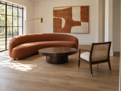 A living room pairing a curved terracotta sofa with a traditional cane-back accent chair, connected by a shared brass lamp finish, beneath a rust-toned abstract textile hanging.