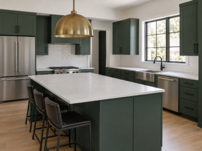A contemporary kitchen with deep green slab cabinetry, a sculptural brass pendant light over the island, matte black cabinet hardware, and a brushed stainless sink.