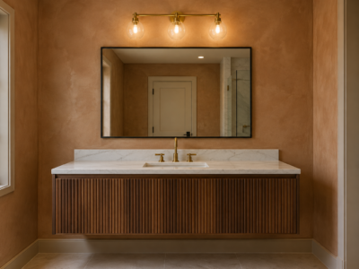 A bathroom vanity with a linear brass bar light mounted above a mirror, a clear gap of wall visible between the mirror and the fixture, against a warm clay-plaster wall.