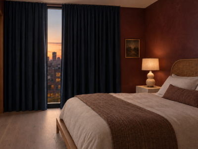 A primary bedroom at dusk with deep ink-navy blackout curtains pulled nearly closed across a full-height window, a low platform bed with a curved cane headboard, and a lit ceramic table lamp on a plinth-form stone nightstand.