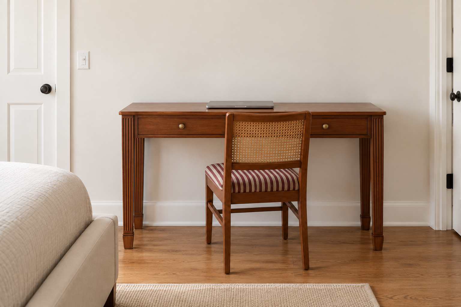 A fluted-leg cherry console table used as a desk in a guest bedroom, with a cane-back chair and a laptop on top.