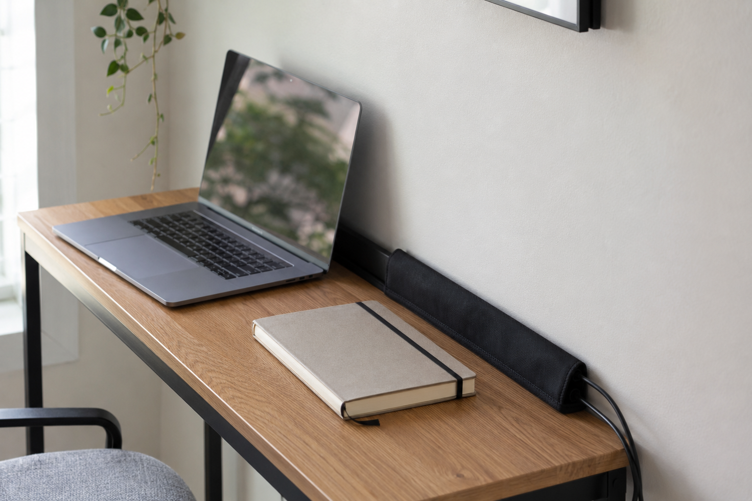 A narrow desk with a laptop and a cord management sleeve along the back edge