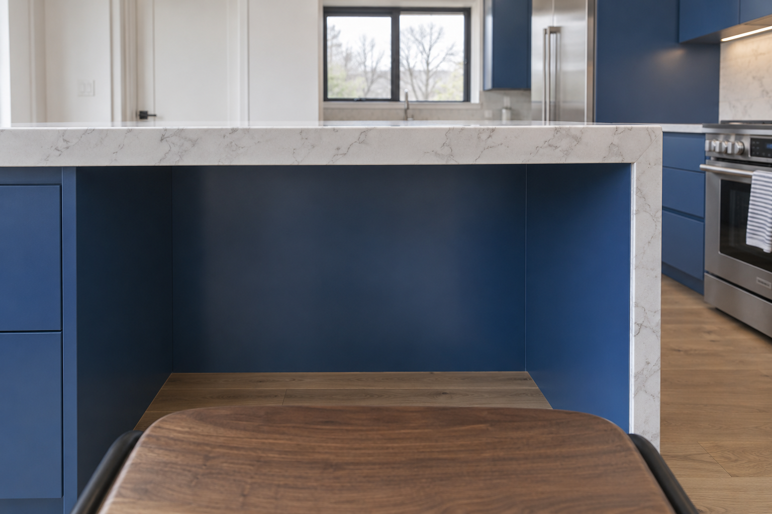 A low, seated-eye-level view showing knee clearance under a cobalt-blue kitchen island's stone counter overhang, beside a walnut-seat counter stool.