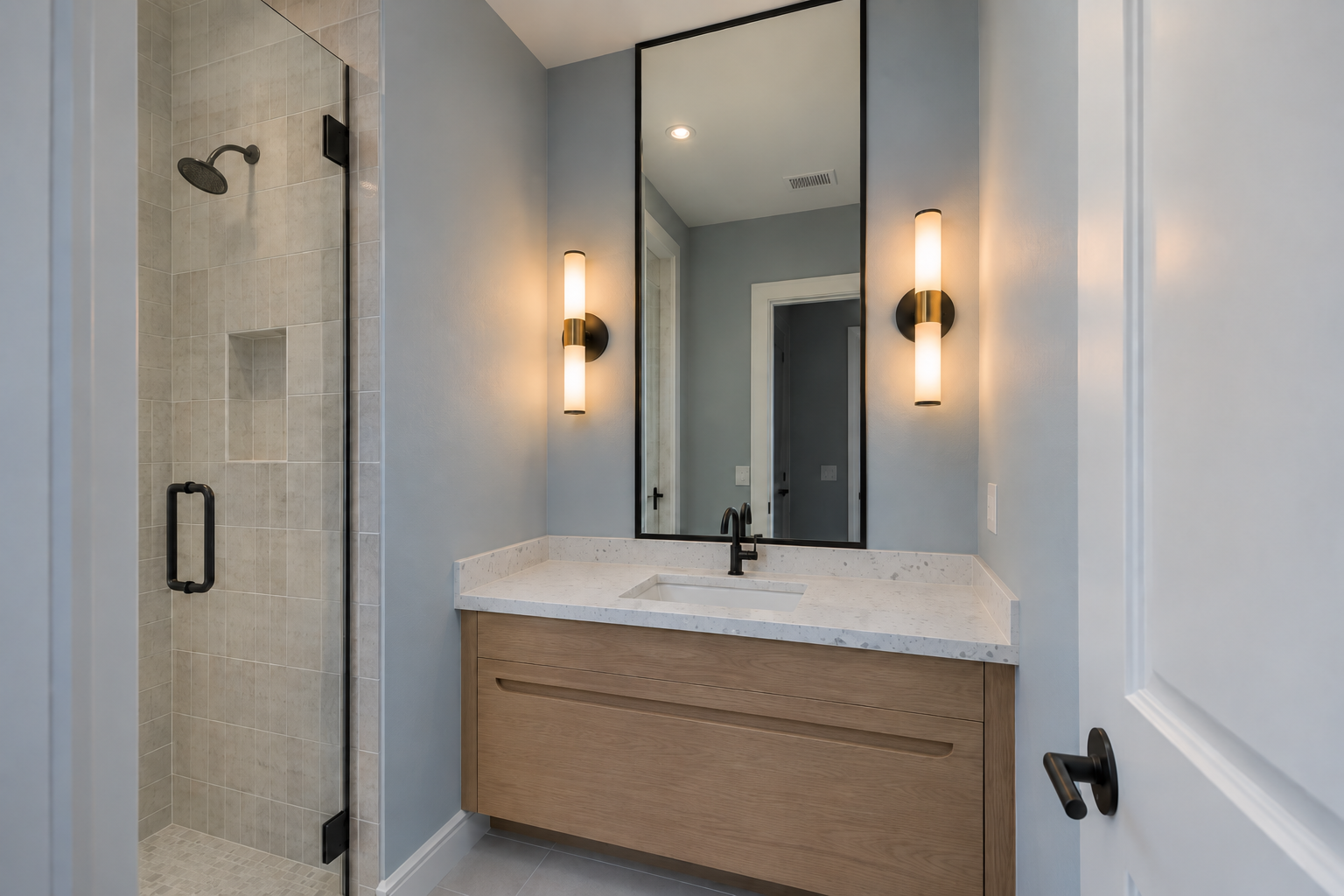 A narrow bathroom vanity wall with a tall mirror reaching close to the ceiling, lit by two black-and-brass cylindrical sconces mounted at eye level on either side instead of a fixture above it.