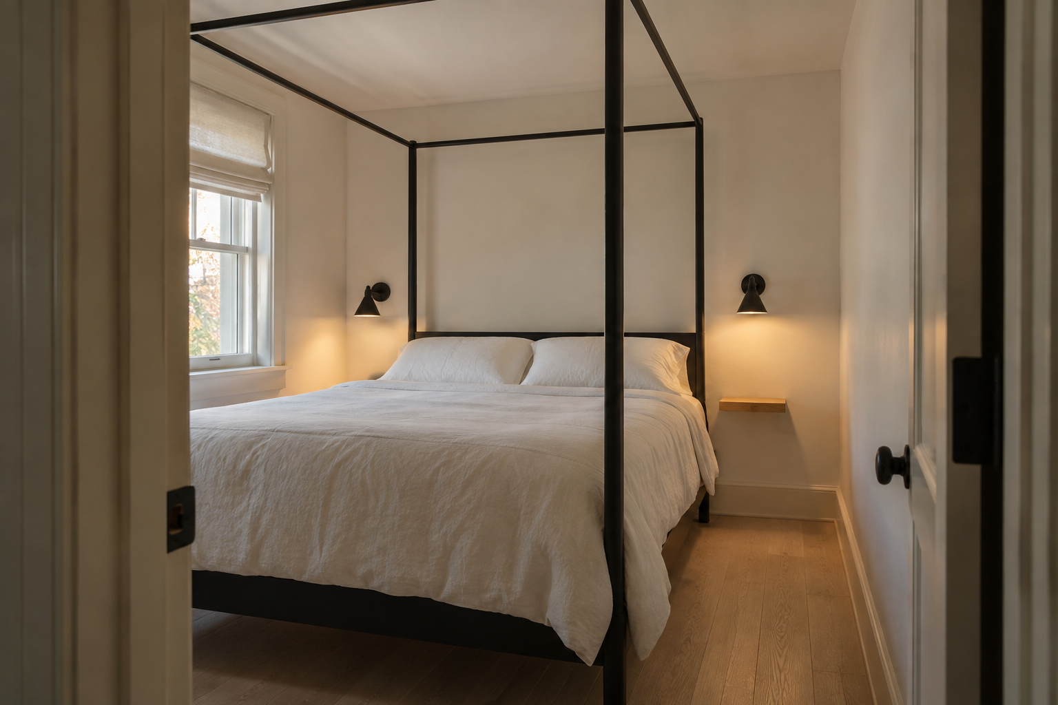 A small bedroom seen from the doorway, with a slim black iron four-poster bed frame and a genuinely narrow walking gap between one post and the side wall.