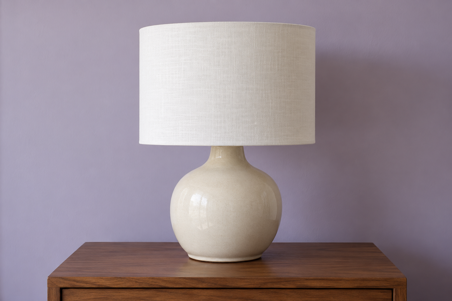 A cream stoneware lamp base with a linen drum shade about two thirds its height, against a lilac wall.