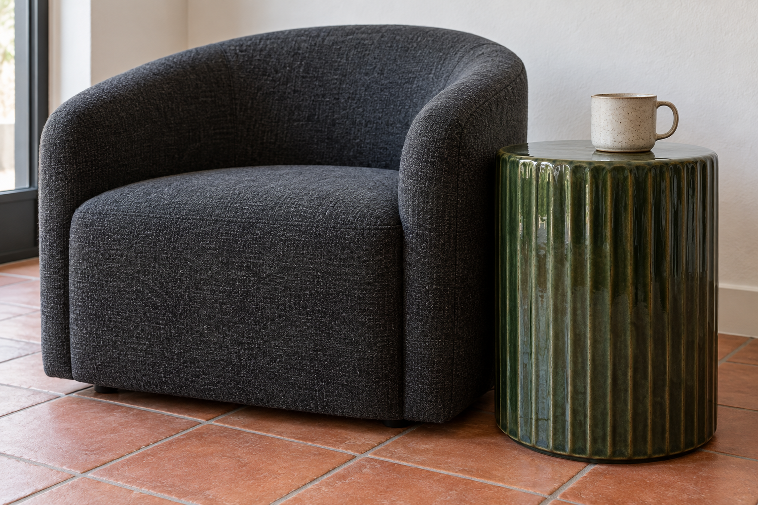 A green ceramic drum side table level with the arm of a charcoal wool armchair, a mug on top.
