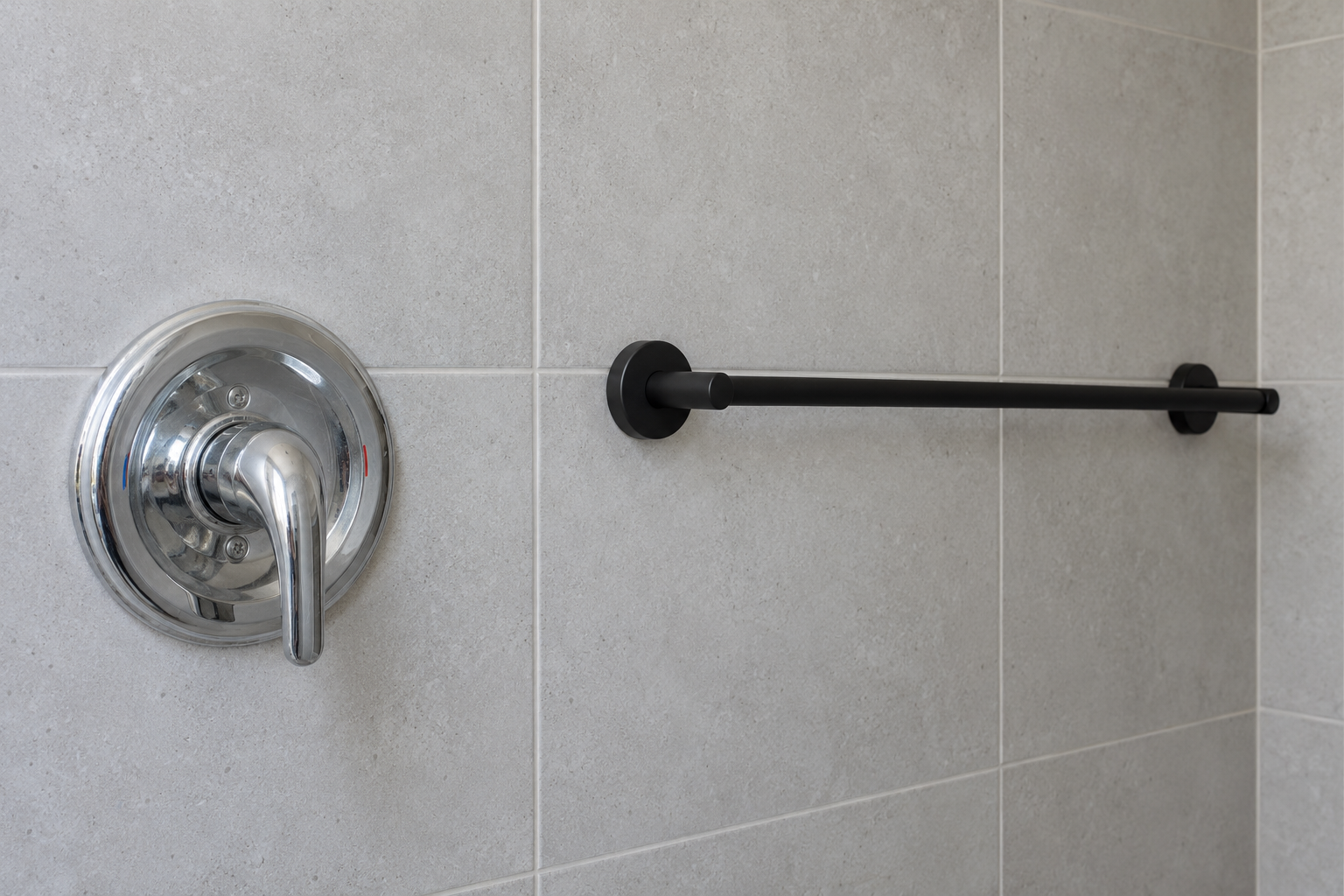 A close detail of an older polished chrome shower valve beside a newer matte black towel bar on a pale gray tiled bathroom wall.