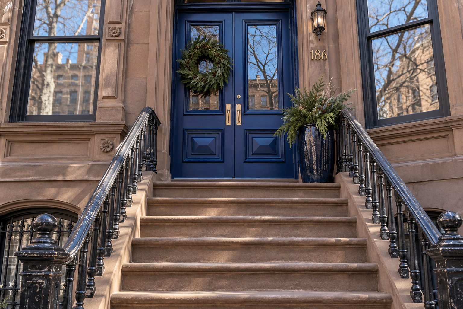 A brownstone stoop with a deep blue door, one wreath, and a single planter of evergreens.