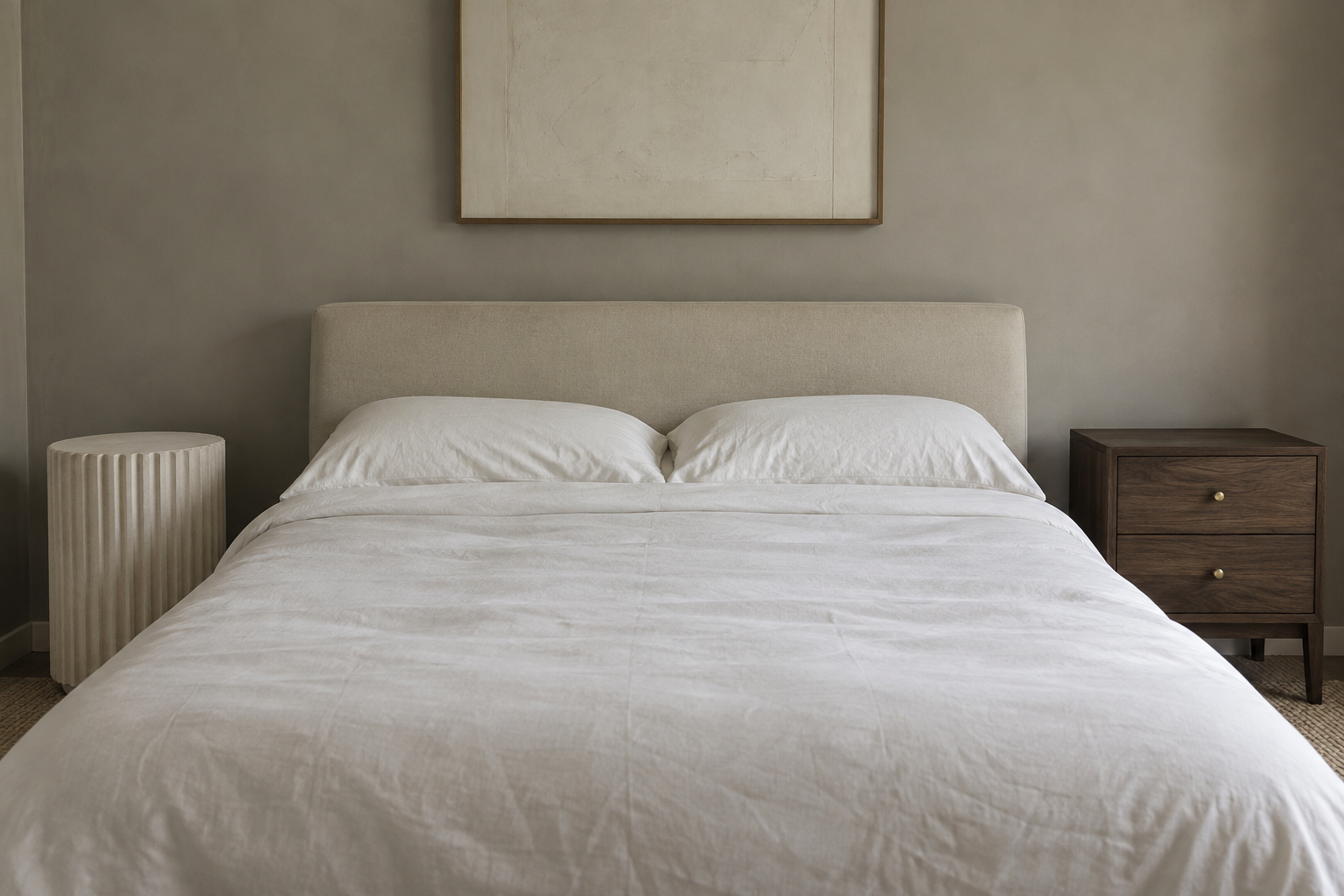 A fluted plaster-toned pedestal nightstand and a small dark-wood chest with brass hardware, seen from bed height on either side of a bed's foot.