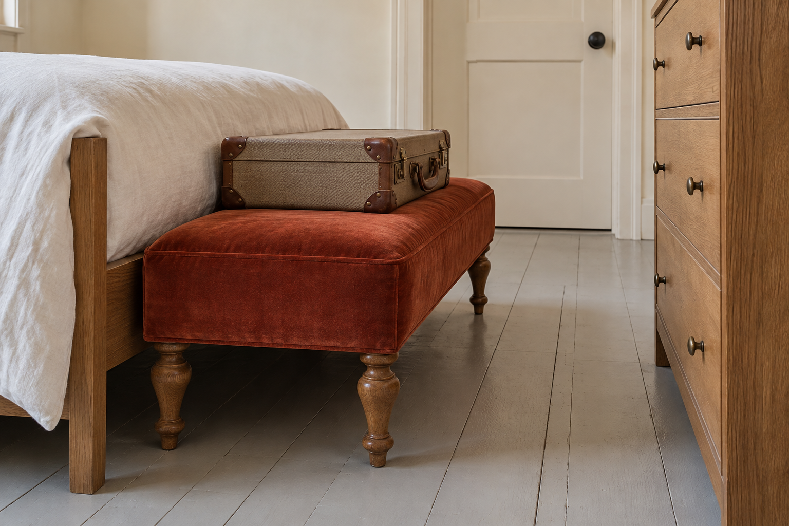 A closed suitcase on a rust velvet bench at the foot of a guest bed.