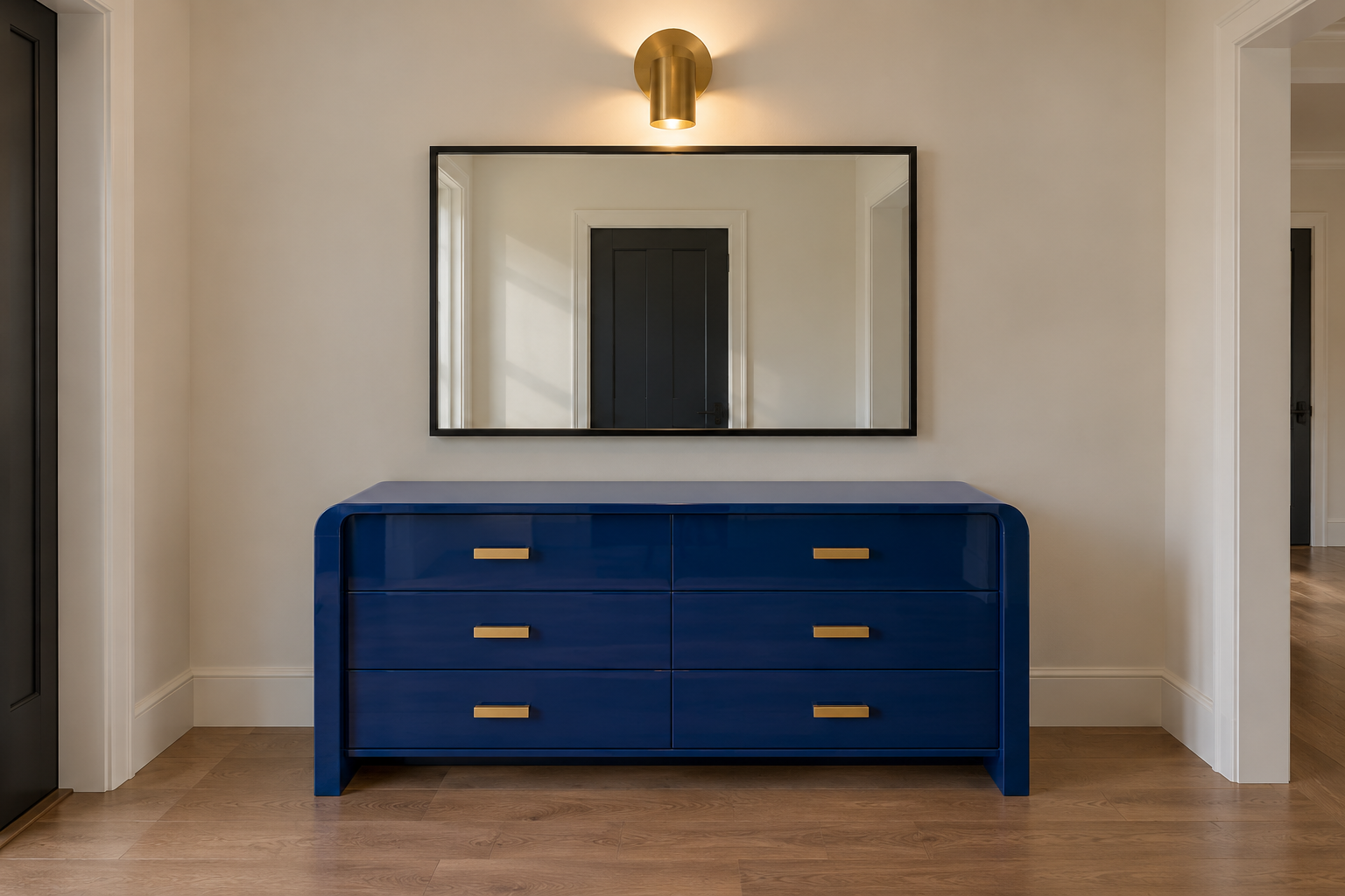 A cobalt lacquered waterfall-edge sideboard with brass drawer pulls, a matte black framed mirror, and a brass wall sconce above it.