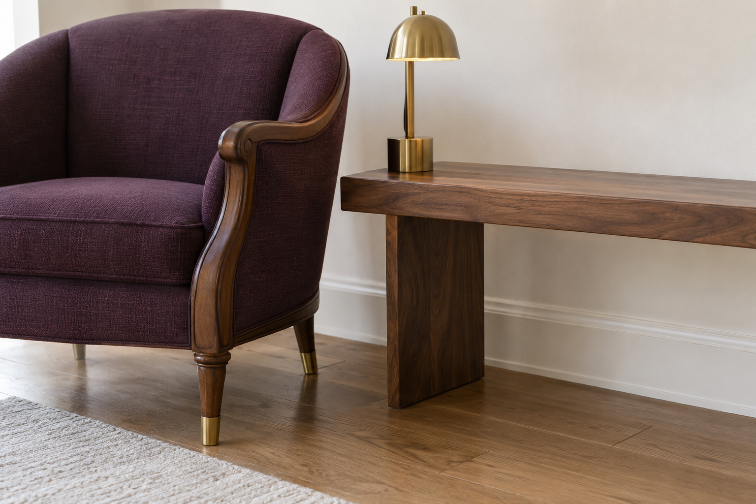 A close view of a traditional plum-upholstered armchair beside a low contemporary walnut bench, connected by a shared brass finish on the chair's feet and a small table lamp.