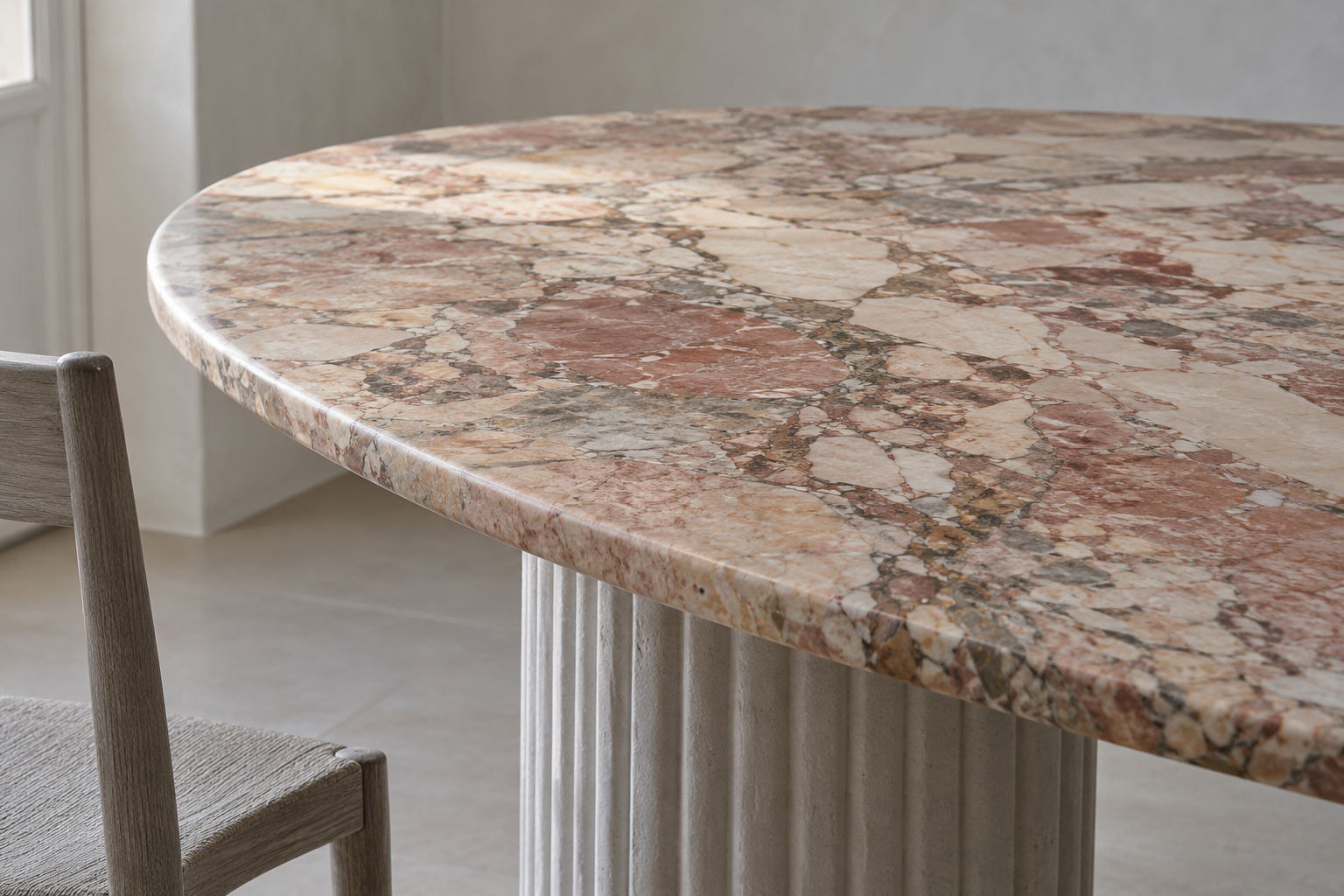 Close view of an oval Breccia marble dining table top with a thick rounded edge above a pale gray fluted base.
