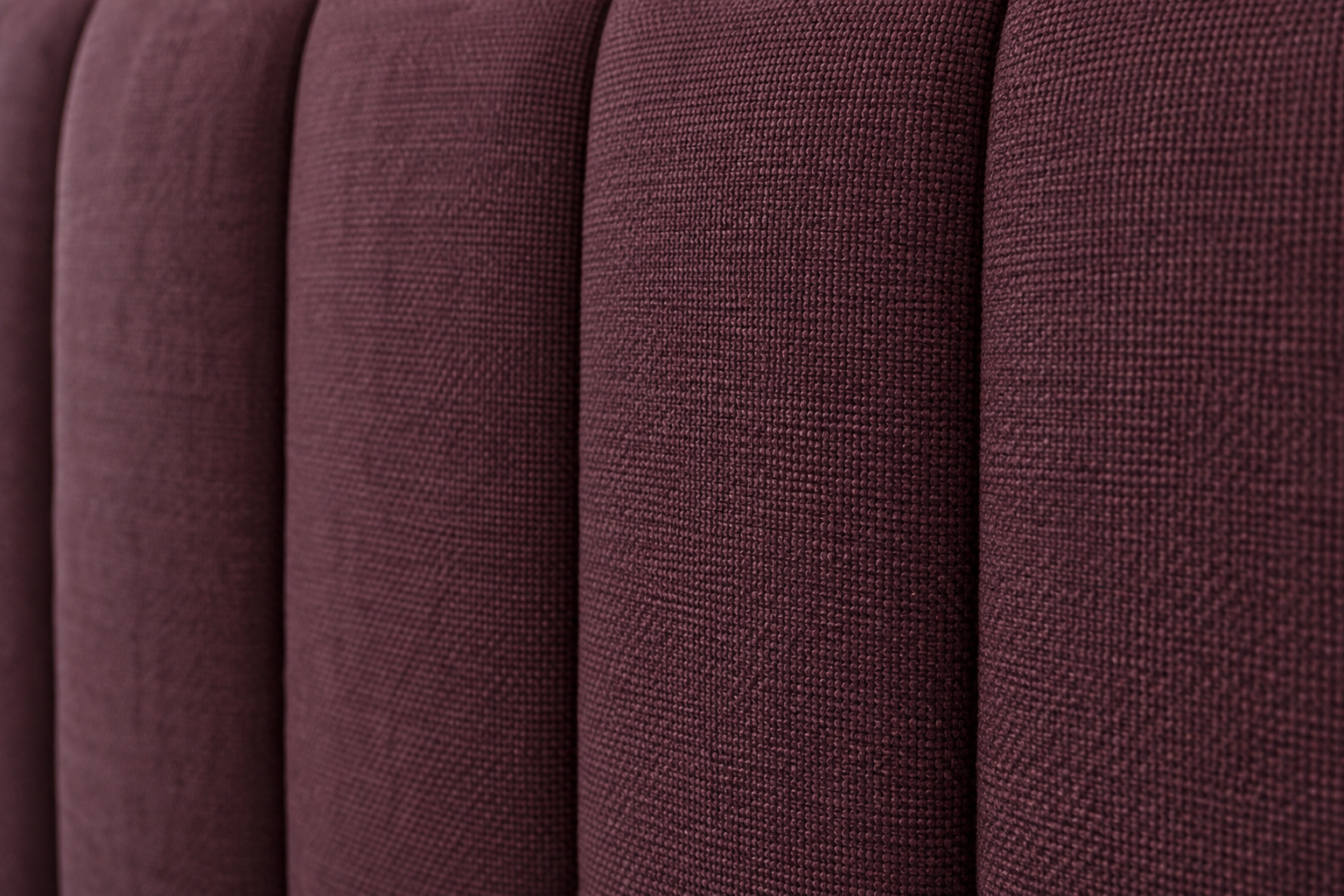 A close detail of a deep aubergine channel-tufted upholstered headboard, raking light revealing the vertical seam construction.