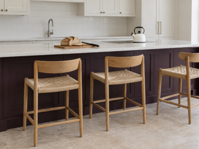 Three oak counter stools with woven seats at a kitchen island with deep aubergine cabinets and a white countertop.