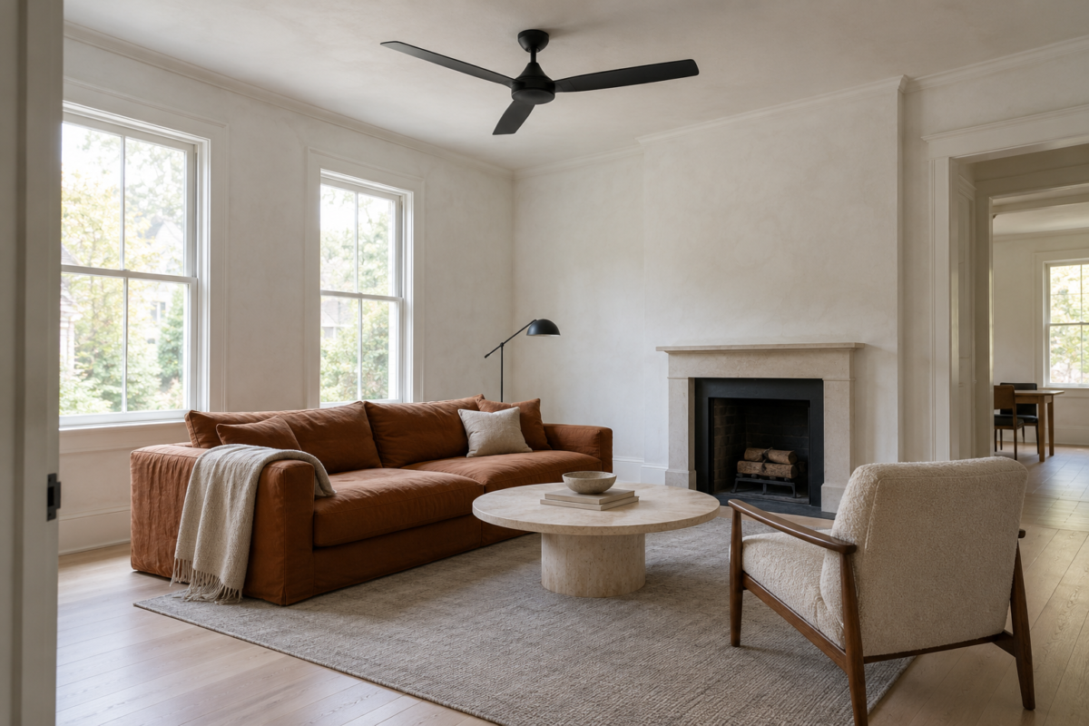 A matte black ceiling fan with three flat blades on a short rod in a white living room with pale ash floors and a low rust linen sofa.