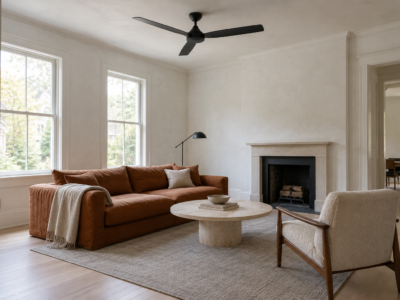 A matte black ceiling fan with three flat blades on a short rod in a white living room with pale ash floors and a low rust linen sofa.