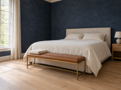 A bedroom with deep ink navy limewashed walls, a queen bed with oatmeal linen bedding, and a caramel leather bench on slim brass legs at its foot, with a folded wool blanket on one end.