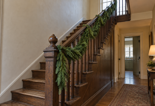 Fresh fir and cedar garland tied along the dark oak banister of a staircase and hanging down at the newel post, with cream plaster walls.