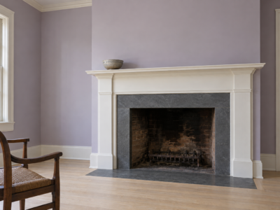 A fireplace with dark soapstone around the opening and a white wood mantel, in a room with lilac walls.