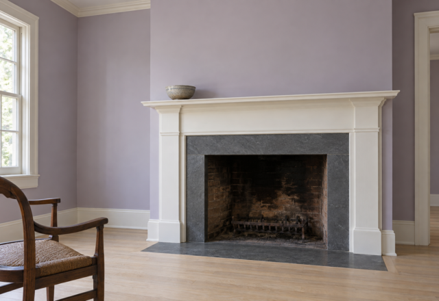 A fireplace with dark soapstone around the opening and a white wood mantel, in a room with lilac walls.