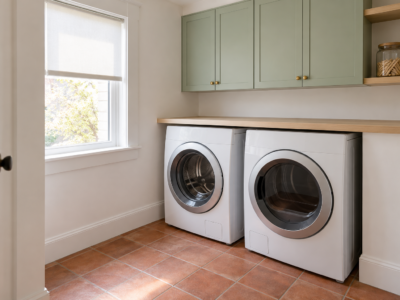 A laundry room with a white front-loading washer and dryer side by side under a pale ash counter, mint green cabinets above, and a terracotta tile floor with clear space in front.