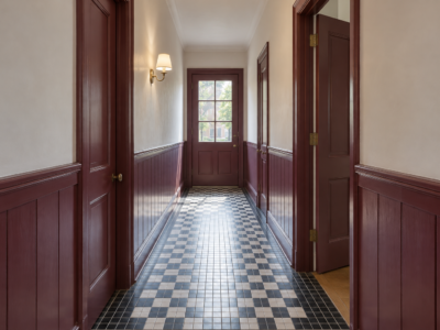 A narrow hallway with mulberry painted paneling below cream walls, a black and white mosaic tile floor, and a glazed door letting in daylight at the far end.