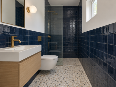 A small bathroom with navy tile to shoulder height, a toilet hung on the wall, a floating oak vanity, and a glass shower at the far end.