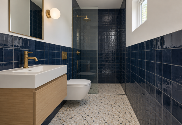 A small bathroom with navy tile to shoulder height, a toilet hung on the wall, a floating oak vanity, and a glass shower at the far end.