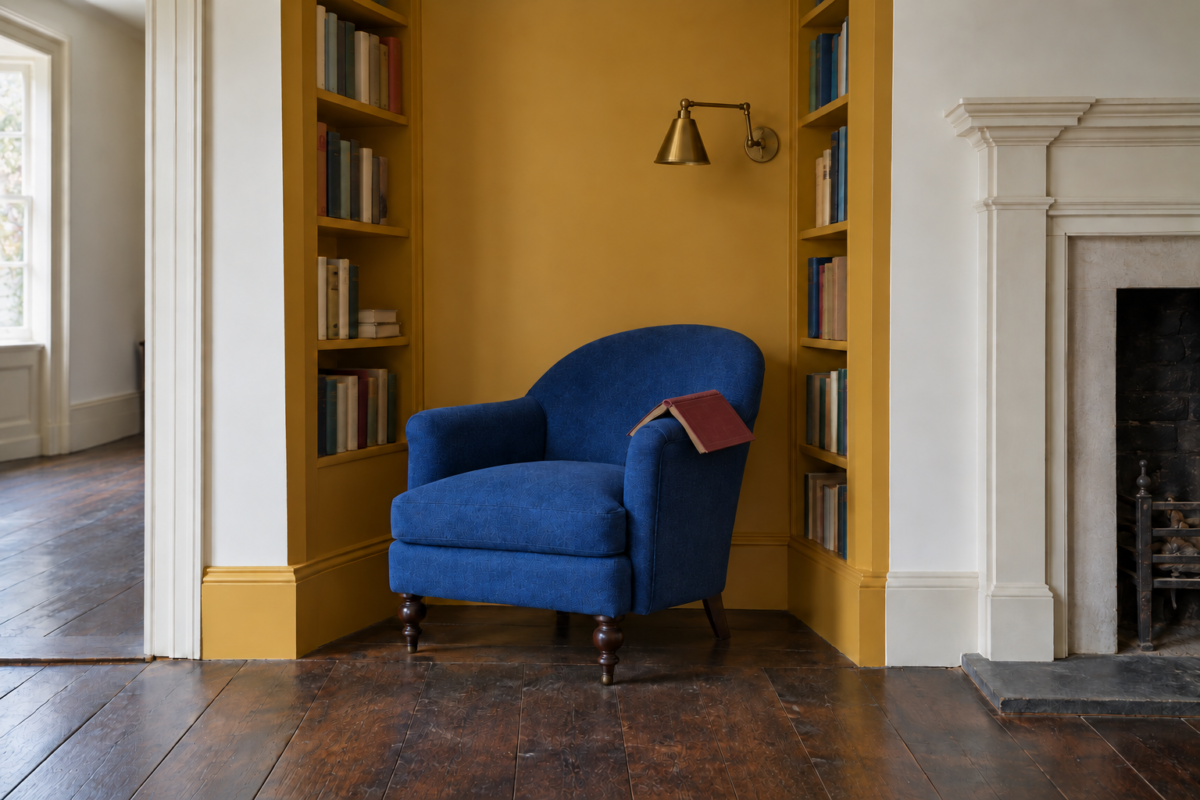 A cobalt blue armchair in an alcove painted ochre yellow, with bookshelves on both sides and a brass wall light above the chair.