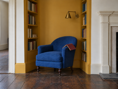A cobalt blue armchair in an alcove painted ochre yellow, with bookshelves on both sides and a brass wall light above the chair.