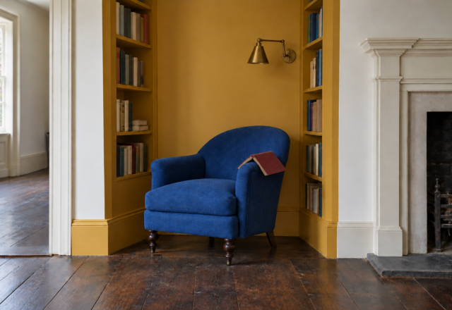A cobalt blue armchair in an alcove painted ochre yellow, with bookshelves on both sides and a brass wall light above the chair.