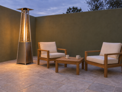 A lit pyramid patio heater with a glass flame tube beside two teak chairs and a small table on a patio with an olive green wall.