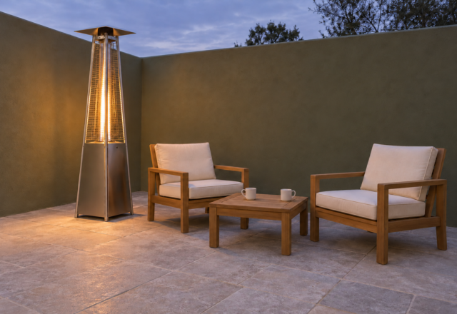 A lit pyramid patio heater with a glass flame tube beside two teak chairs and a small table on a patio with an olive green wall.