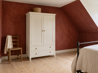 A white painted armoire with shaker doors and brass knobs against a deep red limewashed wall in an attic bedroom, beside an iron bed.