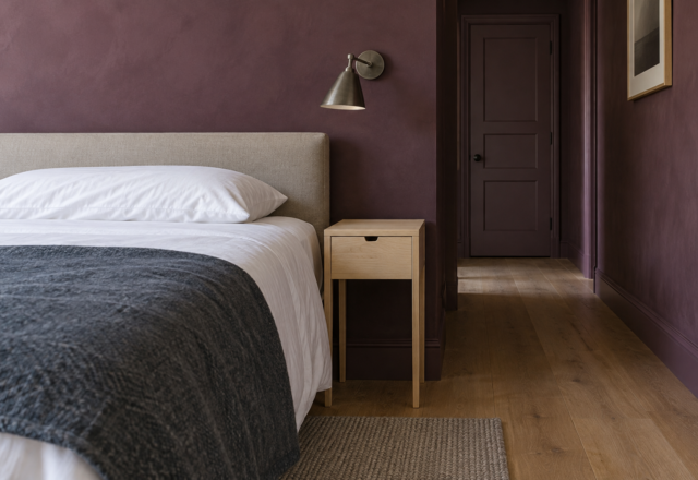 A slim pale maple nightstand level with the mattress beside a white-sheeted bed against a plum limewashed wall, with a pewter wall light above it.