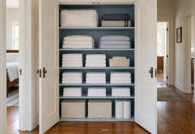 An open linen closet with a powder-blue interior, sheet sets in pillowcases, stacked white towels, and bins on the floor.