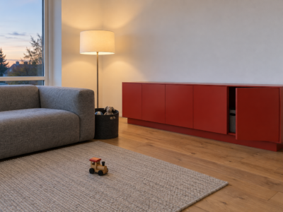 A living room with a long tomato red sideboard, its doors nearly closed, a basket of toys by the sofa, and one toy train on the rug.